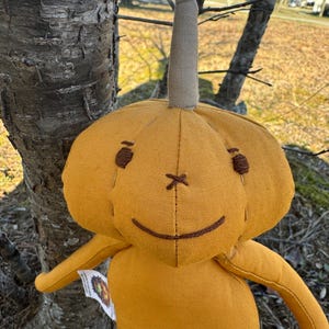 Golden yellow 14-inch Pumpkidz plush doll standing outdoors near a tree, featuring a stitched pumpkin face with a wide gentle smile and cheerful expression. Handmade adoptable pumpkin plush from the Pick-A-Pumpkidz Patch.