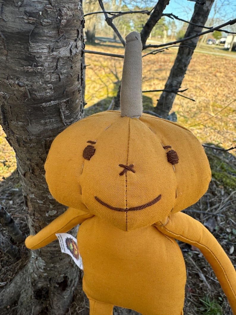 Golden yellow 14-inch Pumpkidz plush doll standing outdoors near a tree, featuring a stitched pumpkin face with a wide gentle smile and cheerful expression. Handmade adoptable pumpkin plush from the Pick-A-Pumpkidz Patch.