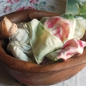 Può includere: Una ciotola di legno vintage culla una bambola di porcellana con i capelli rossi, vestita con un abito bianco. La bambola è adagiata tra sciarpe di seta colorate nei toni del rosa, verde e bianco. Un dipinto è visibile sullo sfondo.