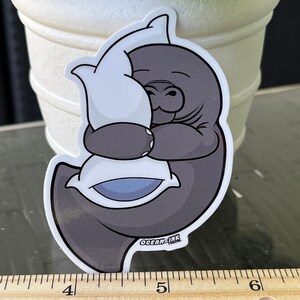 Op de afbeelding: Een grijze cartoon-manatee die een wit kussen met een blauw midden omhelst. De sticker heeft de tekst "OCEAN-INK" onderaan.
