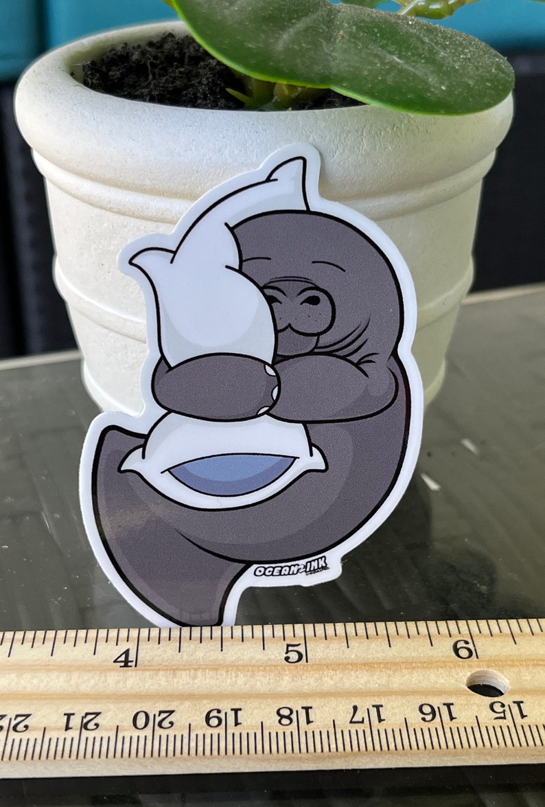 Op de afbeelding: Een grijze cartoon-manatee die een wit kussen met een blauw midden omhelst. De sticker heeft de tekst "OCEAN-INK" onderaan.
