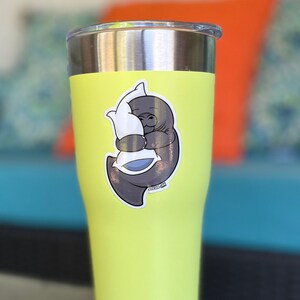 Op de afbeelding: Een gele roestvrijstalen beker met een sticker van een grijze lamantijn die een wit kussen omhelst. De sticker heeft de tekst "Ocean Ink".