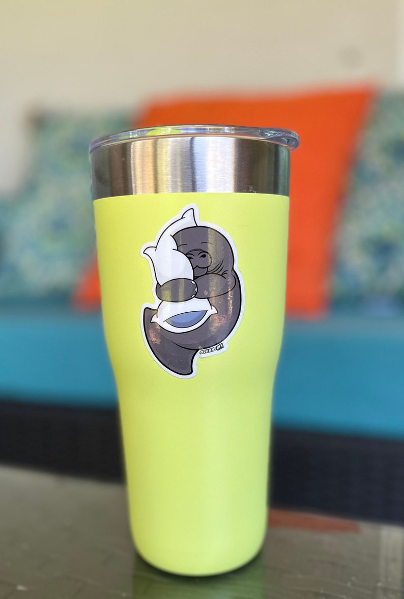 Op de afbeelding: Een gele roestvrijstalen beker met een sticker van een grijze lamantijn die een wit kussen omhelst. De sticker heeft de tekst "Ocean Ink".