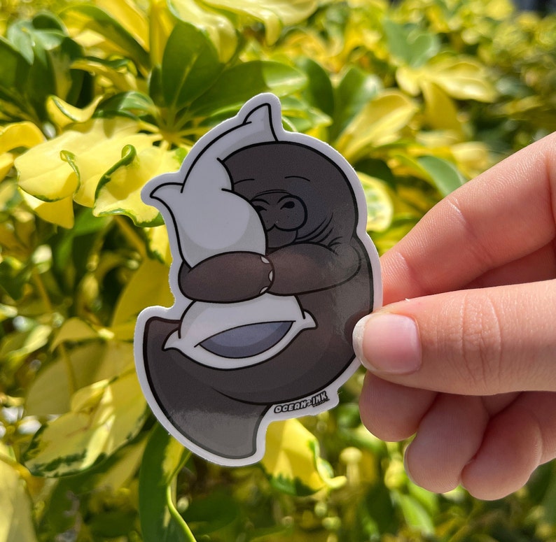 Op de afbeelding: Een sticker van een bruine lamantijn die een wit kussen met blauwe bies omarmt. De sticker heeft de tekst "Ocean Ink" onderaan.