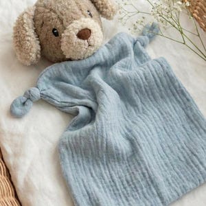 Puede incluir: Un juguete de bebé con forma de perro, con cabeza y orejas de color marrón claro, sobre una manta de seguridad de muselina azul claro. La manta tiene esquinas anudadas y está colocada sobre una superficie blanca dentro de una cesta de mimbre.