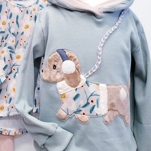Könnte beinhalten: Ein Set Kinderkleidung mit einem hellblauen Kapuzenpullover mit einem braunen und weißen Hundeflicken, einem langärmeligen weißen Shirt mit Blumenmuster und rosa Leggings. Der Pullover hat den Text "Kryezi's mode design" auf der Kapuze.