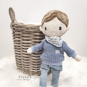 Könnte beinhalten: Eine Plüschpuppe eines Jungen, die einen blauen Pullover mit der Aufschrift "Super Kind", blaue Jeans und einen gestreiften Schal trägt. Die Puppe hält einen schwarz-weißen Fußball und steht auf einer weißen, flauschigen Oberfläche.