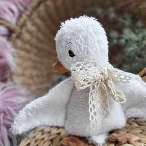 以下が含まれることがあります： 白いぬいぐるみのかわいいアヒル。くちばしは茶色で、足も茶色です。首には白いレースのリボンが結ばれています。アヒルは籐で編まれた表面の上に座っています。