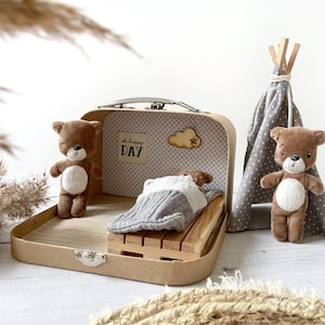 Könnte beinhalten: Ein brauner Koffer mit einem grauen und weißen gemusterten Innenraum. Der Koffer ist geöffnet und hat ein kleines Bett im Inneren, auf dem ein Teddybär schläft. Es gibt ein kleines Holzschild mit der Aufschrift "oh happy DAY" und eine Holzwolke mit einem Stern darauf. Zwei weitere Teddybären stehen außerhalb des Koffers. Ein Teddybär steht neben einem grauen und weißen Tipi.