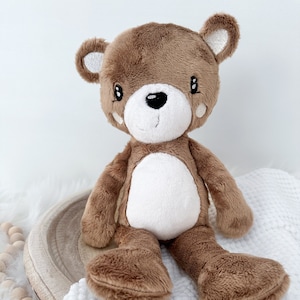 May include: A brown plush teddy bear with white accents on its face and belly. The bear is sitting on a white blanket.
