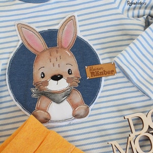 Puede incluir: Una camisa a rayas azules y blancas con una aplicación de conejo marrón. El conejo lleva una bufanda gris y está rodeado de un círculo azul. El texto "kleiner Räuber" está en una etiqueta marrón.