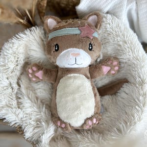 Könnte beinhalten: Ein braunes Plüsch-Katzenspielzeug mit einem hellblauen Stirnband und einem rosa Stern. Die Katze hat einen weißen Bauch, rosa Pfoten und liegt auf einer flauschigen, cremefarbenen Decke. Das Spielzeug befindet sich in einem Weidenkorb.