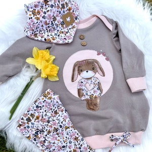 May include: A gray and pink baby onesie with a brown bunny applique. The bunny is wearing a pink dress with a heart and floral pattern. The onesie has a floral print bib and matching leggings.