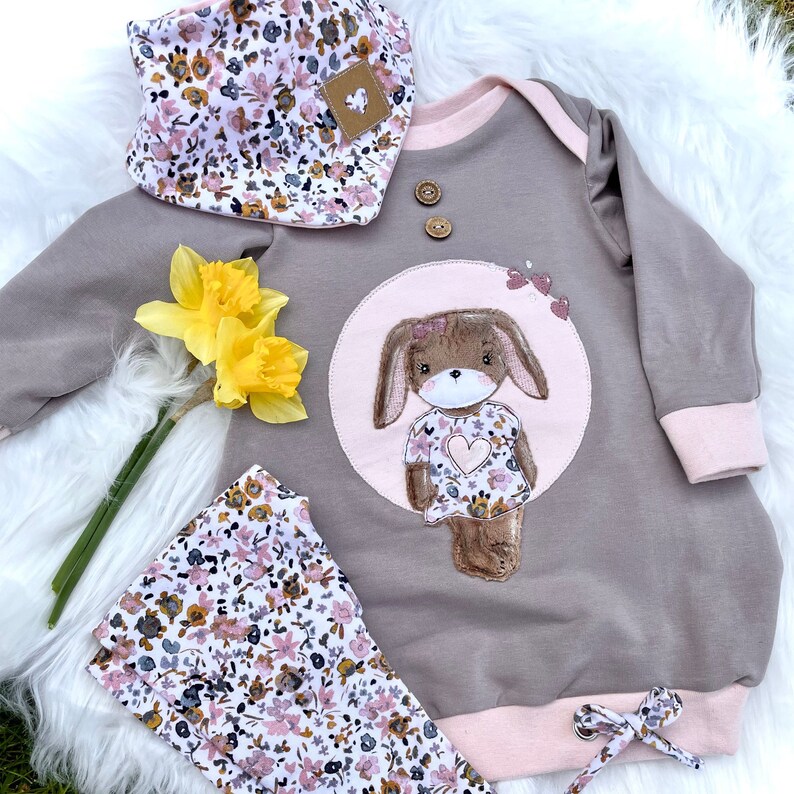 May include: A gray and pink baby onesie with a brown bunny applique. The bunny is wearing a pink dress with a heart and floral pattern. The onesie has a floral print bib and matching leggings.