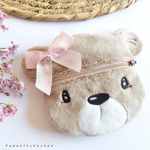 May include: A plush brown bear-shaped zippered pouch with a pink satin bow and a silver heart charm. The pouch has embroidered eyes and a black nose.