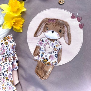 May include: A light brown bunny applique with a pink floral dress and a pink heart on the front. The bunny is sewn onto a light pink circle with a light brown background.