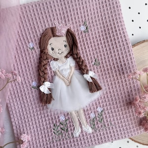 May include: A pink waffle-weave fabric square with an embroidered princess design. The princess has brown plaits, a pink crown, and a white dress. Embroidered flowers and greenery surround the princess.
