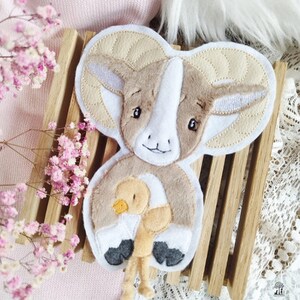 May include: A plush toy of a brown and white ram with large curled horns holding a small yellow chick. The ram is made of soft fabric and has embroidered details.