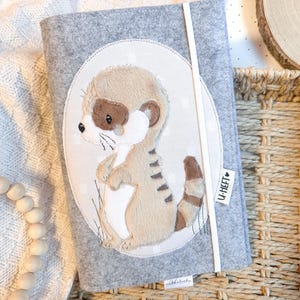 Może przedstawiać: Szara książeczka dla niemowląt z filcu z białym owalem z przodu. Owal przedstawia wyszywanego brązowo-białego suricata z napisem "U-HEFT" w kształcie serca.