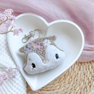 May include: A plush white fox keyring with embroidered brown fur, pink and purple flowers, and black eyes. The keyring is in a white heart-shaped dish.