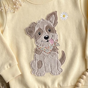 Peut inclure: Un sweat-shirt à manches longues jaune pâle orné d'une grande applique texturée représentant un chiot marron et crème portant un bandana à fleurs. Le chiot tire la langue et une petite marguerite blanche est brodée au-dessus de sa tête.