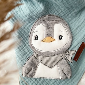 以下が含まれることがあります： グレーと白の刺繍が施されたペンギンがグレーと白のストライプのストールを巻いている、柔らかなブルーのモスリン製ベビーブランケット。ブランケットには、「remarkable」と書かれた茶色のタグが付いています。