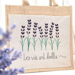 May include: A natural jute tote bag with a white linen panel featuring an embroidered design of lavender flowers and the text "la vie est belle".