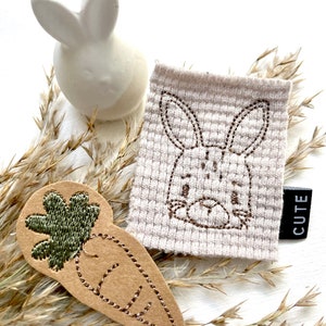 Könnte beinhalten: Eine weiße Keramik-Hasenfigur und ein beiges Stoffquadrat mit einem aufgestickten Hasengesicht. Ein karottenförmiges Applikation mit grünen Blättern und dem Wort "CUTE" auf einem schwarzen Etikett sind ebenfalls vorhanden. Die Artikel sind auf einer weißen Oberfläche mit getrocknetem Gras angeordnet.