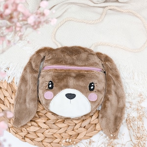 May include: A brown plush bunny-shaped crossbody bag with long floppy ears and a light purple zipper. The bag has a white face with black eyes, a black nose, and pink cheeks. It has a cream-colored rope strap.