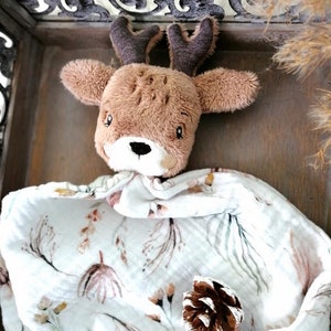 May include: A brown and white deer plush toy with antlers, lying on a white muslin baby blanket with a floral pattern. A brown pine cone is on the blanket.
