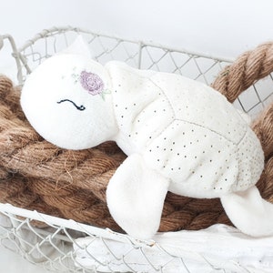 May include: A white plush turtle toy with a pink flower embroidered on its head. The turtle is lying in a white wire basket with brown rope.