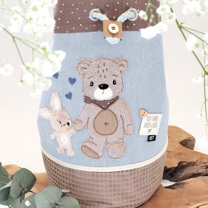 Può includere: Una borsa a cordoncino blu e marrone con un'applicazione di orsacchiotto e coniglietto. La borsa ha il testo "Ich hab dich lieb" e "Hey".