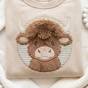 Puede incluir: Sudadera beige con un bordado de una vaca de las Tierras Altas de dibujos animados. La vaca tiene pelaje marrón, cuernos blancos y está dentro de un círculo a rayas. La sudadera es de un material suave.