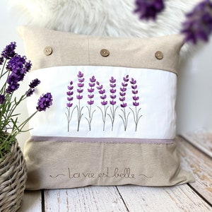 May include: A beige linen pillow with a white panel featuring an embroidered design of purple lavender flowers. The pillow has two beige buttons and the text "la vie est belle" is embroidered on the bottom.