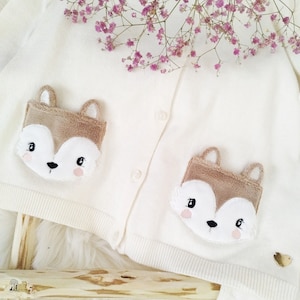 Pode incluir: Cardigan creme com duas aplicações de rosto de raposa. Os rostos das raposas são castanhos, brancos e rosa. O cardigan tem uma frente abotoada e é decorado com um pequeno coração dourado. Um ramo de flores roxas está no fundo.
