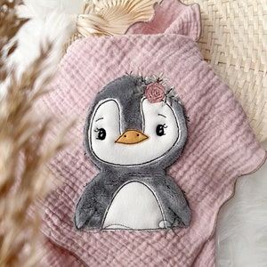 Puede incluir: Una manta de muselina rosa para bebé con un pingüino bordado gris que lleva una corona de flores. La manta tiene un borde festoneado.