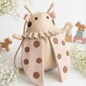 May include: A beige ladybug-shaped backpack with pink wings and brown polka dots. The backpack has a ruffled collar and antennae with pink bows. Wooden toys and flowers are in the background.
