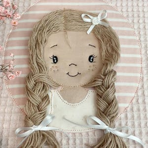 May include: A handmade doll face applique with braided yarn hair, a white top, and a bow. The doll is set against a pink and white striped background, with a pink waffle-textured fabric surrounding it. The doll's face has embroidered details.