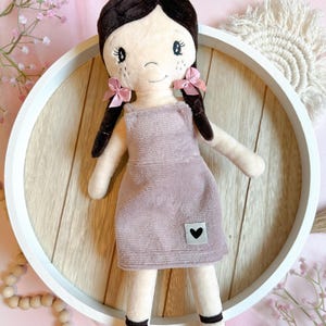 Pode incluir: Uma boneca de pelúcia com cabelo castanho, vestindo um vestido rosa e sapatos pretos. A boneca tem um remendo em forma de coração no vestido.