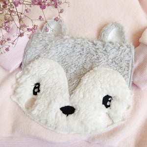 以下が含まれることがあります： ピンクの生地の背景に縫い付けられた、グレーと白のぬいぐるみでできたキツネのアップリケ。黒い目と黒い鼻があります。