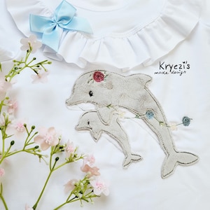 May include: White baby onesie with a gray embroidered design of two dolphins jumping out of the water. The dolphins are surrounded by flowers and the text "Kryezi's mode design" is in the top right corner.