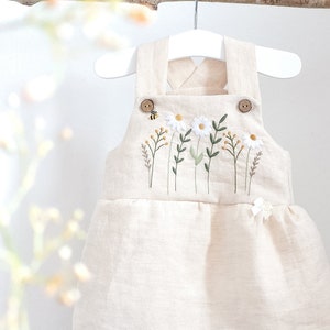 Può includere: Un abito in lino beige con margherite bianche ricamate, foglie verdi e una piccola ape gialla. L'abito ha due fiocchi bianchi sul fondo e due bottoni marroni sulle spalline.