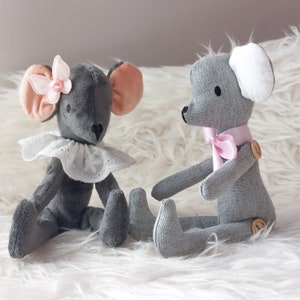 Puede incluir: Dos animales de peluche de tela gris, uno un ratón con un lazo rosa y un cuello de encaje blanco, el otro un oso con un lazo rosa y una oreja blanca.