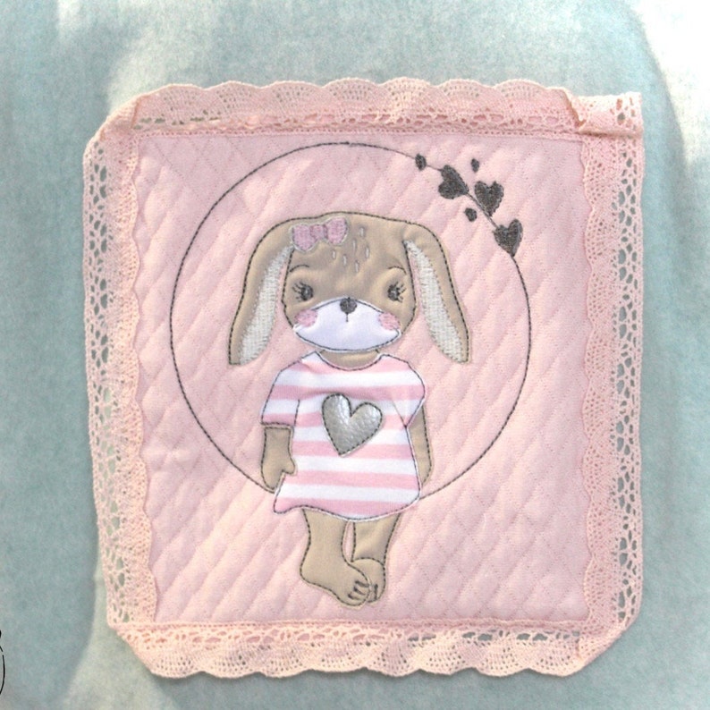 May include: A pink and brown embroidered bunny wearing a pink and white striped dress with a silver heart on the front. The bunny is surrounded by a circle with a vine and hearts.