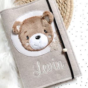 May include: Beige felt baby book cover with a brown bear applique and the name "Levin" embroidered in white thread.