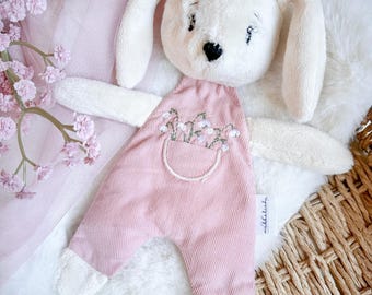 May include: A white plush bunny with long ears wearing pink overalls with embroidered flowers. The overalls have a pocket with a white embroidered outline. The bunny is lying on a white fuzzy surface.