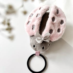 Peut inclure: Un élastique à cheveux en forme de coccinelle, rose et gris. La coccinelle a un extérieur rose duveteux avec des taches grises et une tête grise. Un petit nœud gris est attaché, et un élastique noir est fixé.