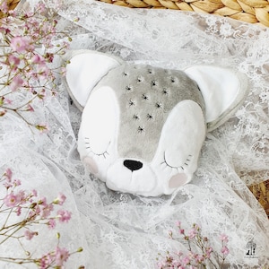 May include: A soft, grey and white deer-shaped cushion with embroidered star details. The cushion has closed eyes, pink cheeks, and a black nose. It rests on white lace, with pink flowers in the foreground.