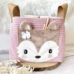 Pode incluir: Saco de armazenamento rosa com textura de waffle com uma aplicação de rosto de raposa marrom e um laço branco. A bolsa tem duas alças de couro marrom.