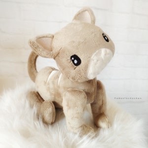 May include: A beige plush toy cat with black eyes and a white nose. The cat is sitting on a white fluffy surface.